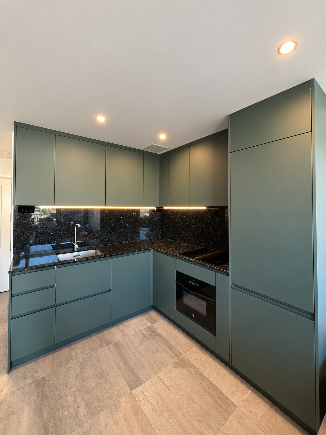 New green L-shaped kitchen with a dark worktop, integrated oven and lighting beneath the wall cabinets